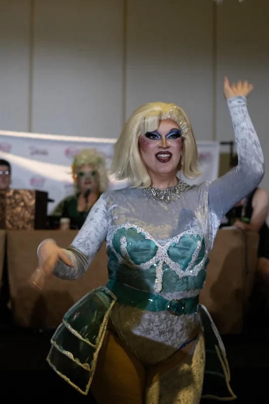A blonde queen in a pale bodice throws one arm in the air.