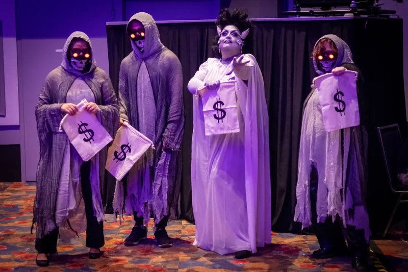 Three NPCs in tattered ghoul robes and glowing skull masks hold out money bags beside @thelolapalooza.